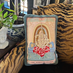 May include: A decorative rectangular pillow with a teal border and a tarot card design. The card features a skeletal hand giving the middle finger, a rainbow, and the text "THE ZERO OF FUCKS". Red roses and blue flowers are at the bottom.