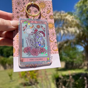 May include: A pink and blue glitter keychain featuring a tarot card with the Lovers card image. The card shows two skeletons holding hands with a red flower between them. The card is surrounded by a gold frame and has the text "THE LOVERS" at the bottom.