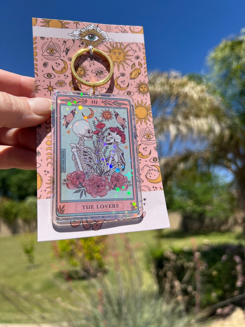 May include: A pink and blue glitter keychain featuring a tarot card with the Lovers card image. The card shows two skeletons holding hands with a red flower between them. The card is surrounded by a gold frame and has the text "THE LOVERS" at the bottom.