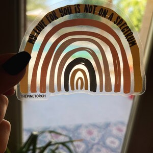 May include: A clear sticker featuring a rainbow design and the text "My love for you is not on a spectrum." The rainbow is made up of various shades of brown, orange, and yellow. The sticker is on a white background.