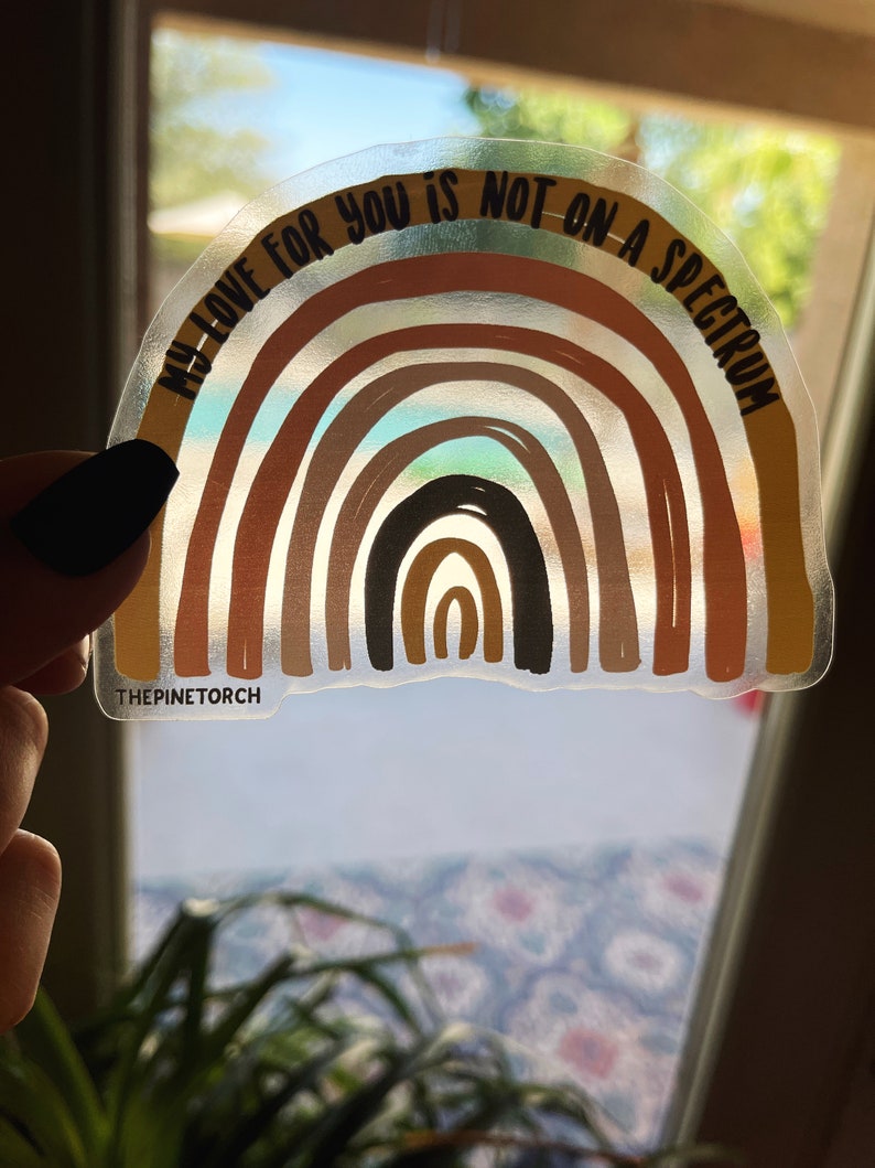 May include: A clear sticker featuring a rainbow design and the text "My love for you is not on a spectrum." The rainbow is made up of various shades of brown, orange, and yellow. The sticker is on a white background.