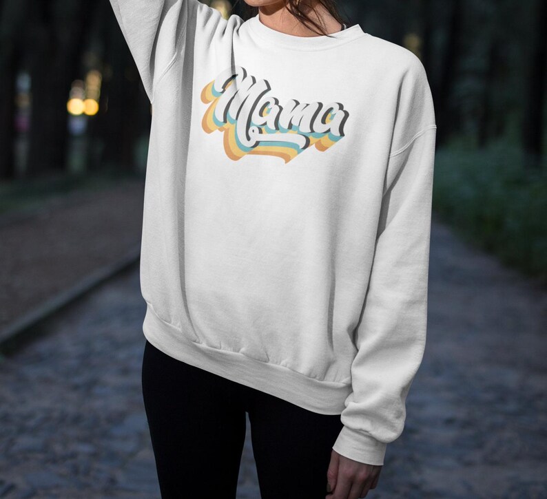 Pode incluir: Camisola branca com um gr&aacute;fico de estilo retro que diz "Mama" em cores do arco-&iacute;ris.