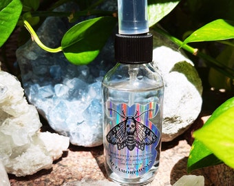 May include: A clear glass bottle with a black spray top and a holographic label. The label features a moth with a skull and the text "CRYSTAL INFUSED SMUDGE MIST" and "METAMORPHOSIS".