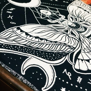 May include: Black and white tapestry featuring a moth with intricate wings and a crescent moon design. The tapestry has a starry background and the text "19" in the bottom right corner.