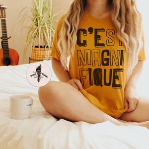May include: A woman wearing a mustard yellow t-shirt with the text "C'EST MAGNIFIQUE" printed in black. The woman is sitting on a white bed with a white coffee mug on the bed next to her.