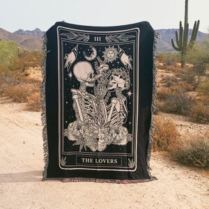 May include: Black woven throw blanket featuring a tarot card design with two skeletons embracing, surrounded by floral elements, celestial symbols, and the text "THE LOVERS". The blanket has a fringed edge.