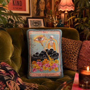 May include: A decorative pillow with a tarot card design featuring a skeleton, rainbow, and eye illustration. The pillow has a light blue background with the words "THE AUDACITY" at the bottom. The pillow is on a green velvet couch.