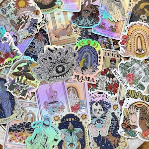 以下が含まれることがあります： ホログラフィックステッカーとタロットカードのコレクション。ステッカーには、目、虹、そして「MAMA」という言葉など、天上的で神秘的なデザインが施されています。占星術のシンボルとイラストが描かれたタロットカードも表示されています。カラーパレットには、紫、青、そして虹色のアクセントが含まれています。