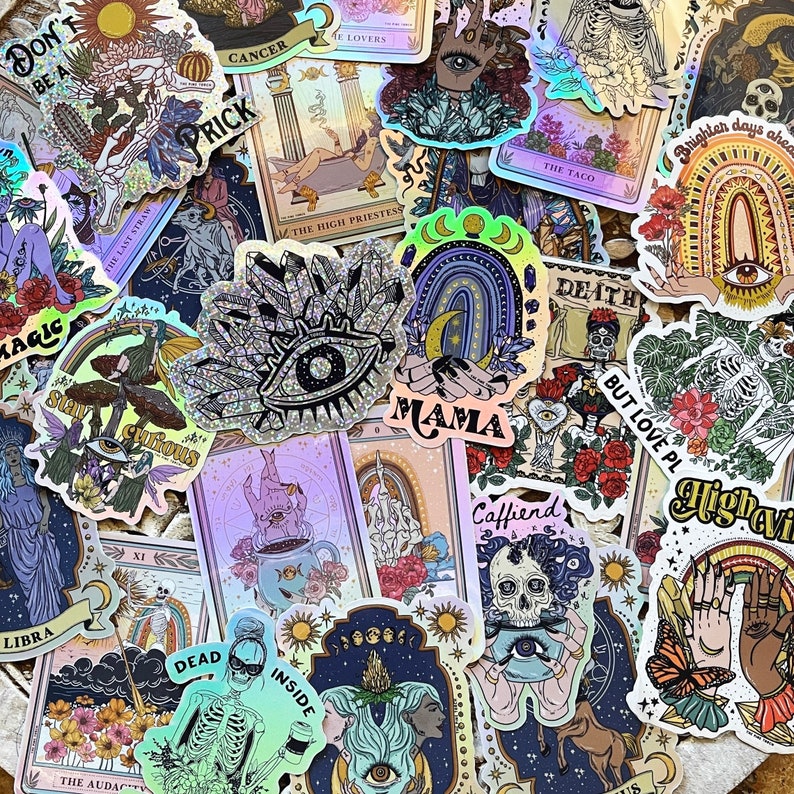 以下が含まれることがあります： ホログラフィックステッカーとタロットカードのコレクション。ステッカーには、目、虹、そして「MAMA」という言葉など、天上的で神秘的なデザインが施されています。占星術のシンボルとイラストが描かれたタロットカードも表示されています。カラーパレットには、紫、青、そして虹色のアクセントが含まれています。