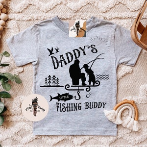 May include: A light gray t-shirt with a black graphic that reads "Daddy's Little Fishing Buddy." The design features a silhouette of a father and child fishing, a fish, trees, and birds. The shirt has short sleeves.