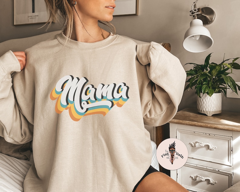 Pode incluir: Uma camisa de moletom bege com um gr&aacute;fico de estilo retr&ocirc; que diz "Mama" em letras coloridas e onduladas.