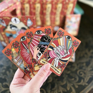 May include: Four tarot cards with colorful illustrations and ornate frames. The cards are held in a hand and feature images of a skull with a hair stylist's scissors, a skeleton hand holding a bottle of shampoo, and a skull with a hair dryer. The cards are from the "The Last Unicorn Tarot" deck.
