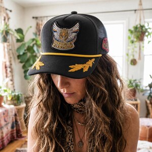Puede incluir: Una gorra tipo camionero negra con parte trasera de malla y un cordón amarillo en la visera. El frente muestra un parche bordado de un leopardo con alas y el texto "SPONSORED BY MY ANCESTORS", con bordados de hojas amarillas en la visera.