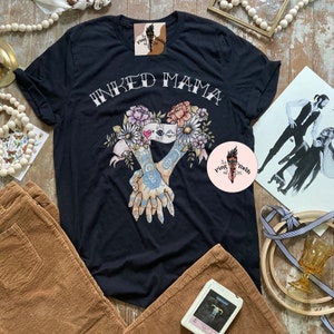 Pu&ograve; includere: T-shirt nero con la scritta bianca "INKED MAMA". La maglietta presenta un disegno floreale con due mani intrecciate, decorate con tatuaggi. Il disegno include fiori rosa, pesca e blu.