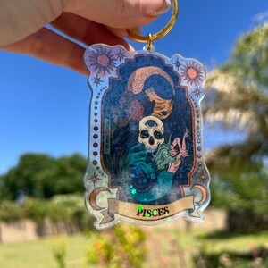 Puede incluir: Un llavero de pl&aacute;stico transparente con un anillo dorado. El llavero presenta una ilustraci&oacute;n colorida de una sirena con una calavera y el texto "Pisces".