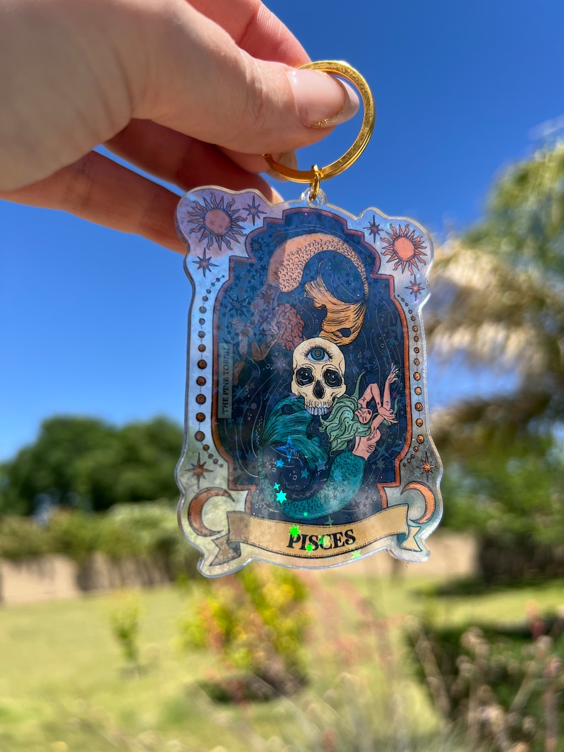Puede incluir: Un llavero de pl&aacute;stico transparente con un anillo dorado. El llavero presenta una ilustraci&oacute;n colorida de una sirena con una calavera y el texto "Pisces".