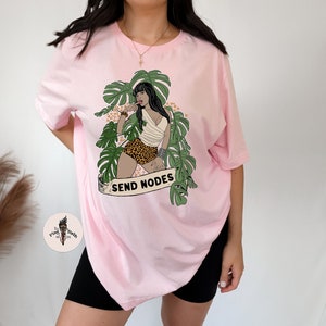 K&ouml;nnte beinhalten: Rosa, &uuml;bergro&szlig;es T-Shirt mit einem Aufdruck einer Frau in einer Dschungelkulisse mit dem Text "SEND NODES".