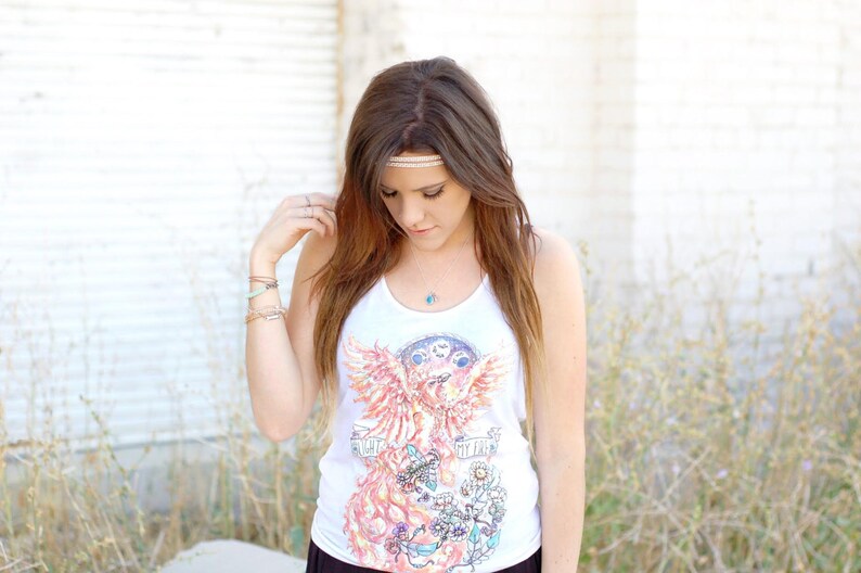 May include: A woman with long brown hair wearing a white tank top with a phoenix graphic and the text "Give Fire" on the front. She is wearing a beaded headband and several bracelets on her wrist.