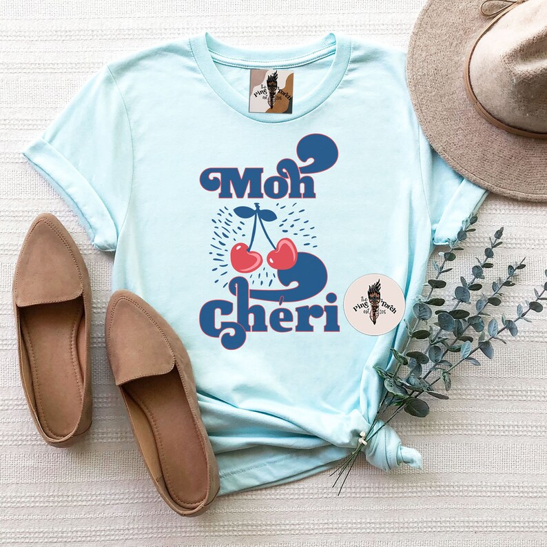 May include: A light blue t-shirt with a retro-style graphic of two red cherries and the text "Mon Cheri" in blue.