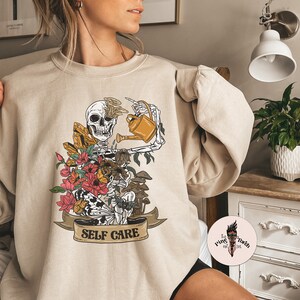 May include: A beige sweatshirt with a graphic print of a skeleton watering flowers and mushrooms. The graphic includes the text "SELF CARE".
