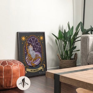 May include: A framed print of a zodiac sign, Scorpio, with a woman sitting on a scorpion. The print has a dark background with gold and purple accents. The text "Scorpio" is written in a banner at the bottom of the print.