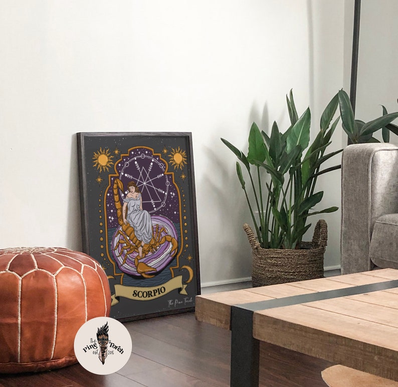 May include: A framed print of a zodiac sign, Scorpio, with a woman sitting on a scorpion. The print has a dark background with gold and purple accents. The text "Scorpio" is written in a banner at the bottom of the print.