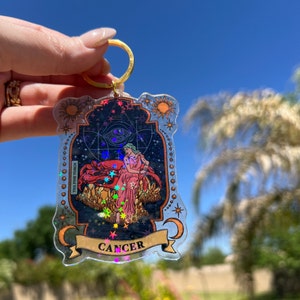 May include: A holographic keychain featuring the astrological sign Cancer. The keychain is decorated with stars, a crescent moon, and a woman sitting on a throne. The text "Cancer" is printed on a banner at the bottom of the keychain.