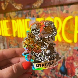 May include: A holographic sticker featuring a skeleton watering flowers with a watering can. The skeleton has a skull with a floral design. The sticker has the text "SELF CARE" and "THE PINE TORCH" on a banner.
