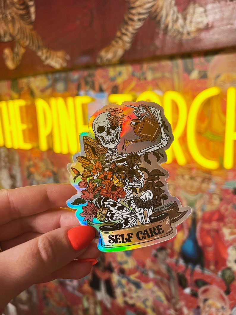 May include: A holographic sticker featuring a skeleton watering flowers with a watering can. The skeleton has a skull with a floral design. The sticker has the text "SELF CARE" and "THE PINE TORCH" on a banner.