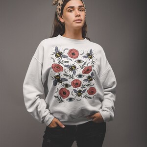 K&ouml;nnte beinhalten: Wei&szlig;er Pullover mit einem Blumendruck mit roten Mohnblumen, Bienen und Augen. Der Druck wiederholt sich auf dem gesamten Pullover.