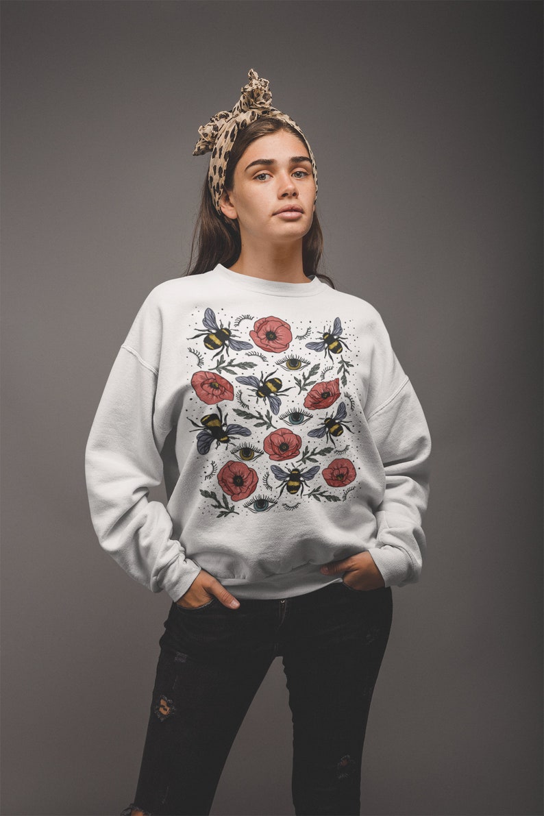 K&ouml;nnte beinhalten: Wei&szlig;er Pullover mit einem Blumendruck mit roten Mohnblumen, Bienen und Augen. Der Druck wiederholt sich auf dem gesamten Pullover.