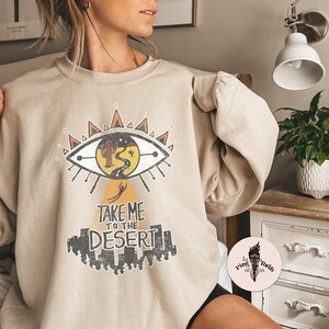 May include: A beige sweatshirt with a graphic print of an eye with a desert scene inside. The text "Take Me To The Desert" is printed below the eye.