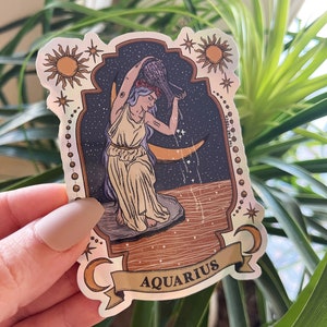 以下が含まれることがあります： ホログラフィックステッカー。水瓶座の星座のイラストが特徴です。デザインには、星空と三日月を背景に、水瓶から水を注ぐ女性が描かれています。「AQUARIUS」の文字がバナーに表示されています。