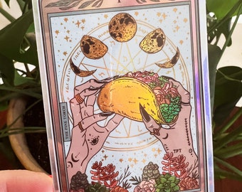 May include: A holographic tarot card with a gold border and a depiction of a hand holding a taco. The card is titled "The Taco" and features a crescent moon, stars, and the text "The Pine Torch".