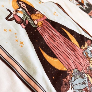May include: A tarot card illustration featuring a woman with long dark hair holding a baby and a cup. The woman is surrounded by stars, moons, and a snake. The card is framed in a peach and blue border.