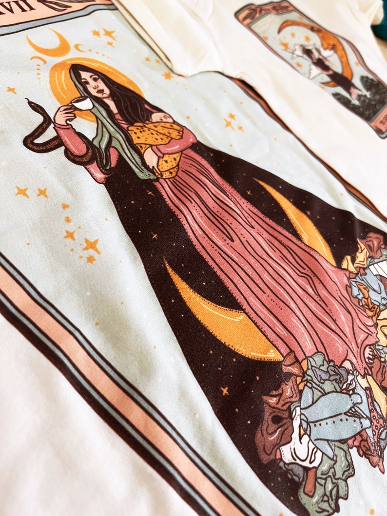 May include: A tarot card illustration featuring a woman with long dark hair holding a baby and a cup. The woman is surrounded by stars, moons, and a snake. The card is framed in a peach and blue border.