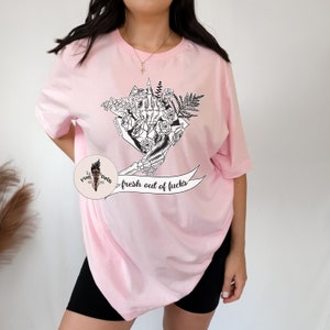 K&ouml;nnte beinhalten: Hellrosa T-Shirt mit einer schwarz-wei&szlig;en Grafik einer Skeletthand, die einen Blumenstrau&szlig; h&auml;lt, mit dem Mittelfinger ausgestreckt. Der Text "fresh out of fucks" befindet sich auf einem Banner unter der Grafik.