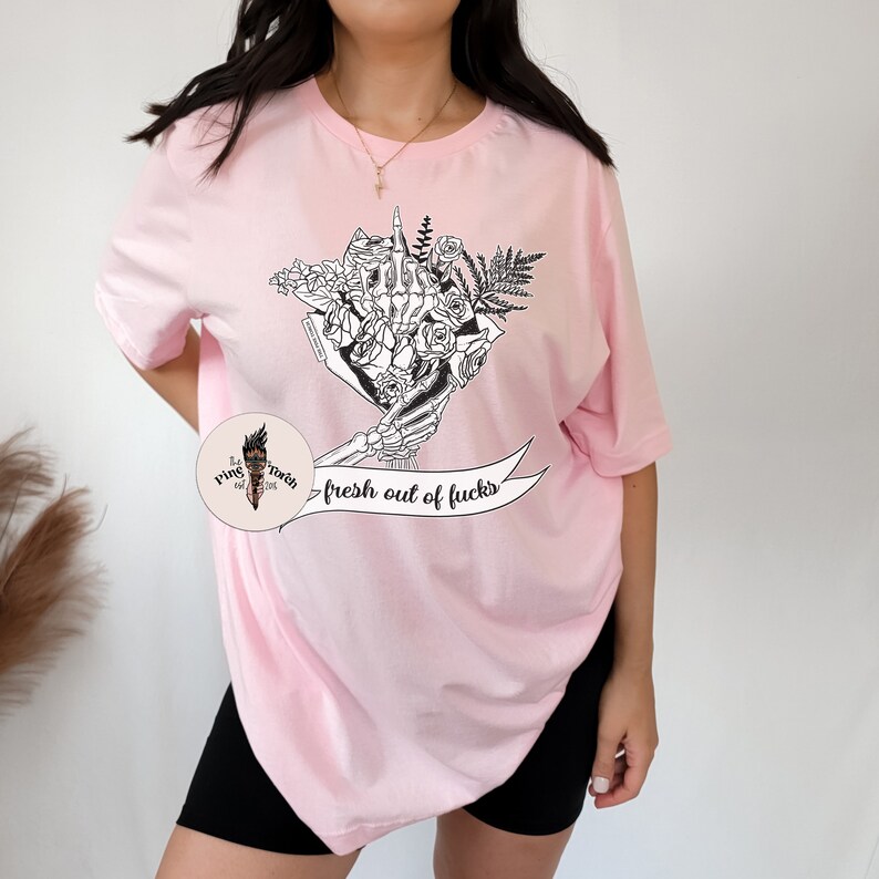 K&ouml;nnte beinhalten: Hellrosa T-Shirt mit einer schwarz-wei&szlig;en Grafik einer Skeletthand, die einen Blumenstrau&szlig; h&auml;lt, mit dem Mittelfinger ausgestreckt. Der Text "fresh out of fucks" befindet sich auf einem Banner unter der Grafik.