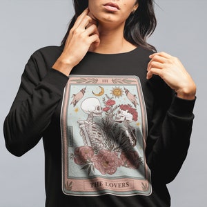 May include: Black sweatshirt with a graphic of a tarot card depicting two skeletons embracing. The card is titled "The Lovers" and has the Roman numeral "III" in the top right corner.