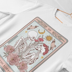 May include: A white sweatshirt with a graphic print of a tarot card. The card is titled "The Lovers" and features two skeletons holding hands with flowers and celestial symbols around them.