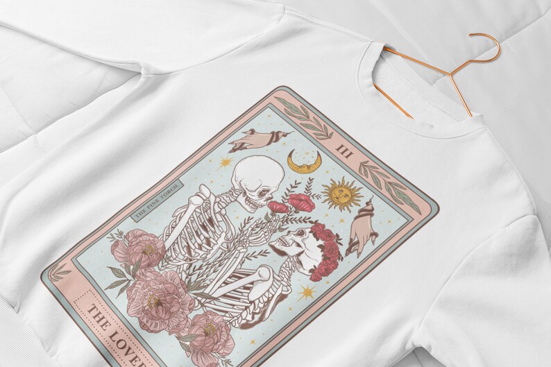 May include: A white sweatshirt with a graphic print of a tarot card. The card is titled "The Lovers" and features two skeletons holding hands with flowers and celestial symbols around them.