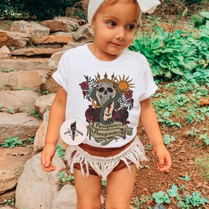 May include: A white t-shirt featuring a graphic of a skull wearing a crown, surrounded by pomegranates and a snake. The text on the shirt reads "Persephone's Pomegranates".