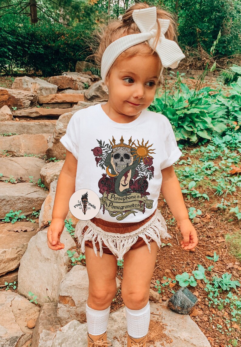 May include: A white t-shirt featuring a graphic of a skull wearing a crown, surrounded by pomegranates and a snake. The text on the shirt reads "Persephone's Pomegranates".