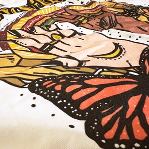 May include: A white fabric with a colourful butterfly design. The butterfly has black wings with orange and red accents. The fabric also features a hand with a crescent moon design.