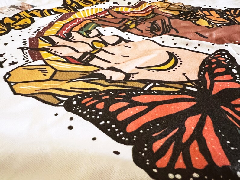 May include: A white fabric with a colourful butterfly design. The butterfly has black wings with orange and red accents. The fabric also features a hand with a crescent moon design.