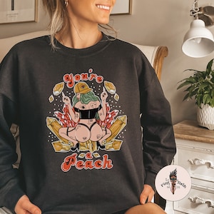 May include: Black sweatshirt with a graphic print of a woman sitting on crystals. The text "You're a Peach" is printed in a retro font.