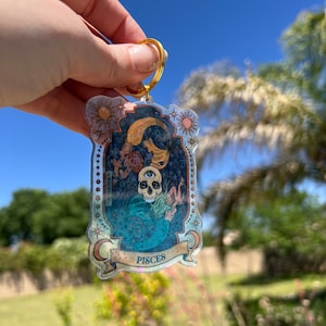 Puede incluir: Un llavero dorado con un dije de acr&iacute;lico transparente que presenta una ilustraci&oacute;n colorida de una calavera con un tercer ojo, una sirena y estrellas. El dije tiene la palabra "Pisces" escrita en una pancarta en la parte inferior.