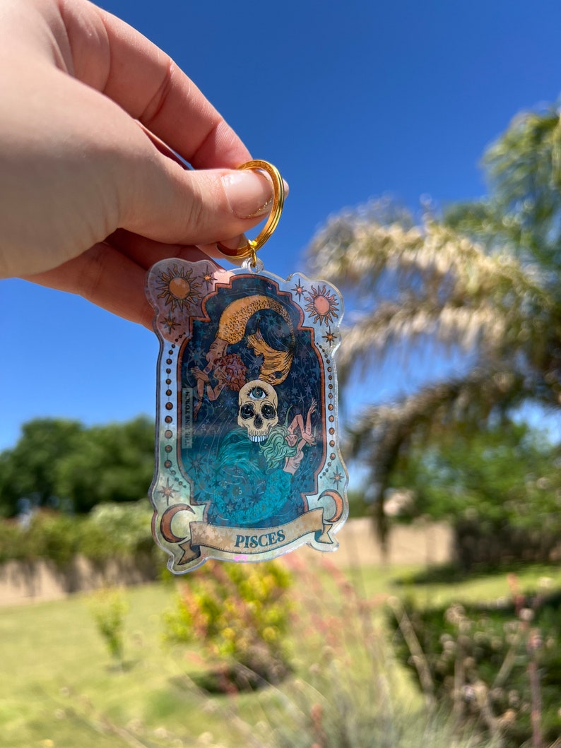 Puede incluir: Un llavero dorado con un dije de acr&iacute;lico transparente que presenta una ilustraci&oacute;n colorida de una calavera con un tercer ojo, una sirena y estrellas. El dije tiene la palabra "Pisces" escrita en una pancarta en la parte inferior.