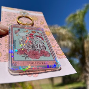 May include: A clear keychain with a holographic finish features a tarot card design. The card depicts two skeletons holding hands in front of a floral background. The card reads "THE LOVERS".
