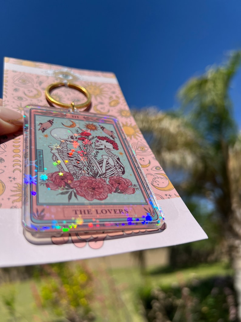 May include: A clear keychain with a holographic finish features a tarot card design. The card depicts two skeletons holding hands in front of a floral background. The card reads "THE LOVERS".